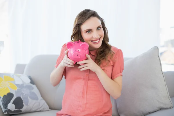 Smiling pregnant woman shaking pink piggy bank sitting on couch — Stock Photo, Image