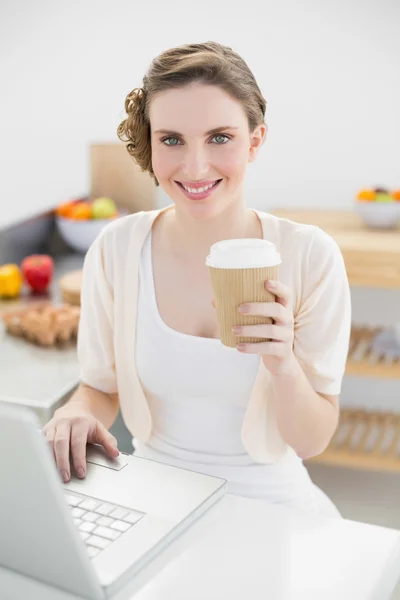 Preciosa mujer usando su cuaderno en su cocina sosteniendo una taza desechable —  Fotos de Stock