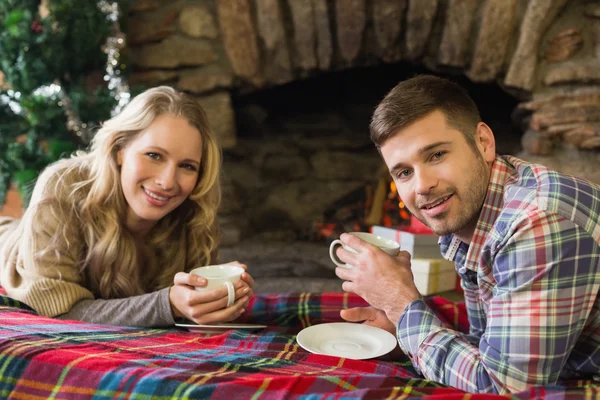 Pareja sonriente con tazas de té frente a la chimenea encendida — Foto de Stock