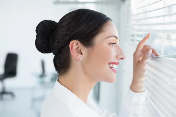 Lachende jonge zakenvrouw gluren via blinds op kantoor — Stockfoto