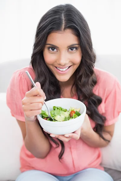 Fröhliche süße Brünette sitzt auf der Couch mit Salatschüssel — Stockfoto