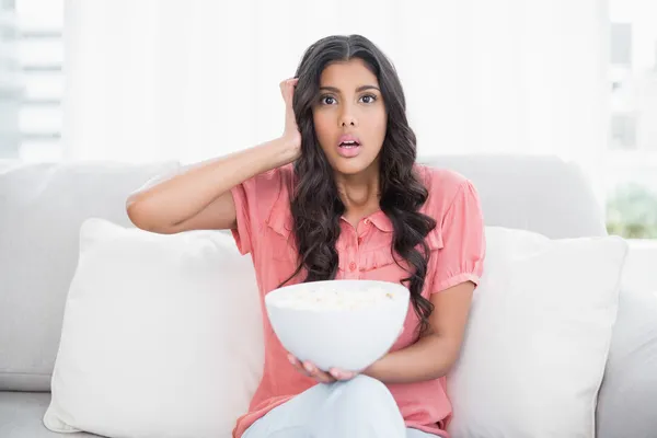 Schockierte süße Brünette sitzt auf Couch und hält Popcorn-Schüssel — Stockfoto