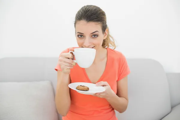 Söt brunett kvinna att lukta en kopp som sitter på soffan — Stockfoto
