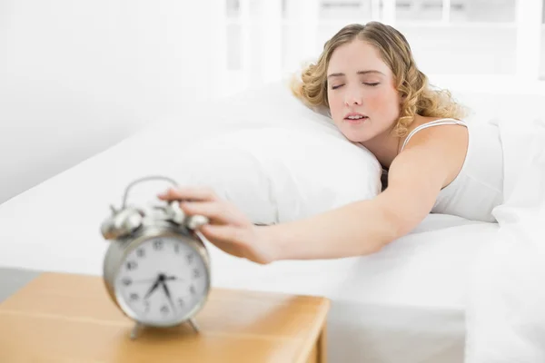 Bastante cansada rubia acostada en la cama alcanzando el despertador —  Fotos de Stock