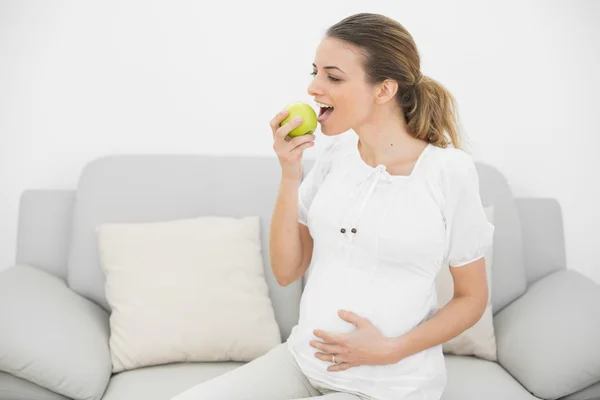 Schöne schwangere Frau isst grünen Apfel, während sie ihre Glocke berührt — Stockfoto