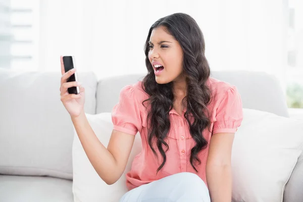 Angry cute brunette sitting on couch holding smartphone — Stock Photo, Image