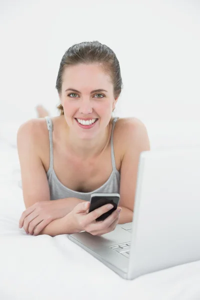 Свободная женщина с мобильным телефоном и ноутбуком в постели — стоковое фото