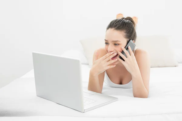 Casual woman using cellphone and laptop in bed — Stock Photo, Image