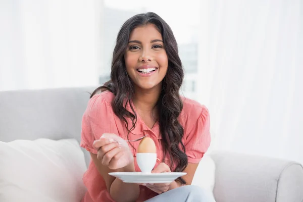 Vrolijke schattige brunette zittend op de bank holding hard gekookt ei — Stockfoto