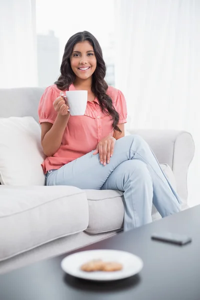 Ler söt brunett sitter på soffan håller mugg — Stockfoto