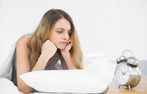 Задумчивая брюнетка лежит под одеялом на кровати
