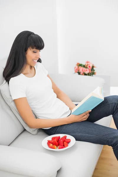 Innehåll mörka haired kvinna läser en bok medan du sitter på soffan — Stockfoto