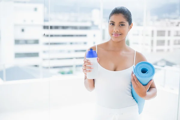 Conteúdo morena tonificada segurando garrafa de esportes e tapete de exercício — Fotografia de Stock