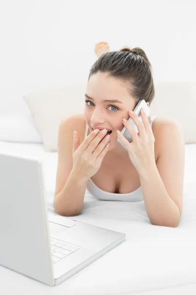 Shocked woman using cellphone and laptop in bed — Stock Photo, Image