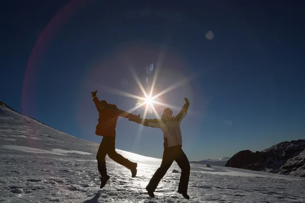 Casal de silhueta pulando na neve contra o sol e o céu azul — Fotografia de Stock