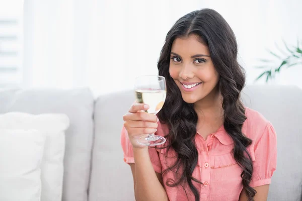 Smiling cute brunette sitting on couch holding white wine glass — Stock Photo, Image