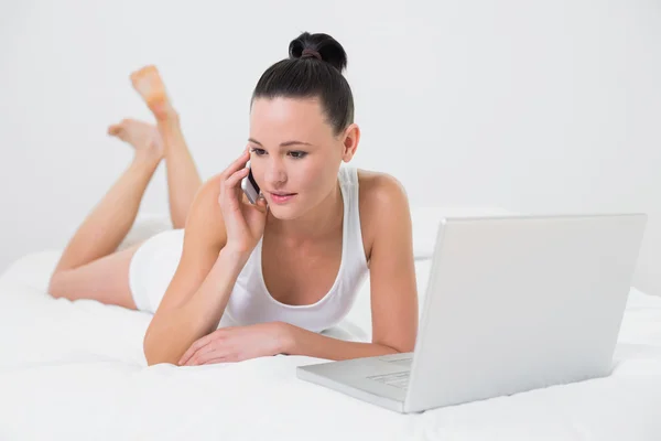 Casual woman using cellphone and laptop in bed — Stock Photo, Image