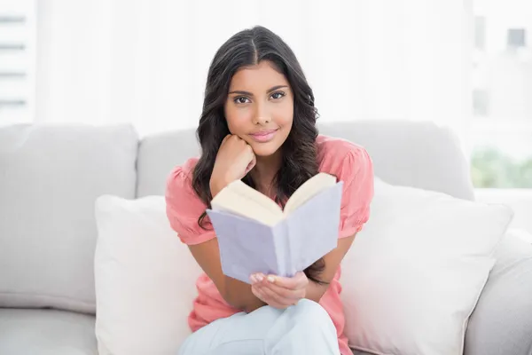 Glad söt brunett sitter på soffan läser en bok — Stockfoto