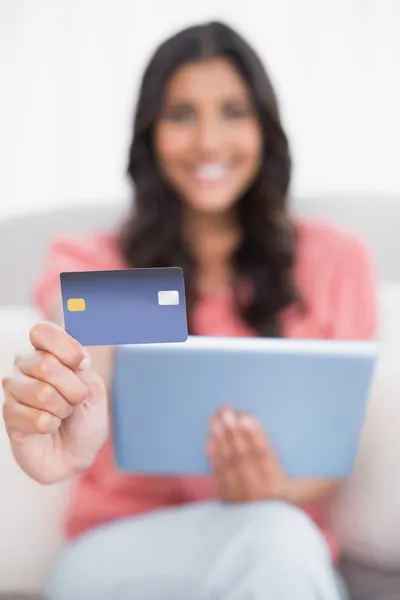 Šťastný roztomilý bruneta sedí na gauči, ukazují kreditních karet držení tabletu — Stock fotografie