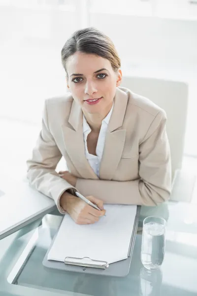 Softly smiling businesswoman writing on a clipboard looking at camera — Stock Photo, Image