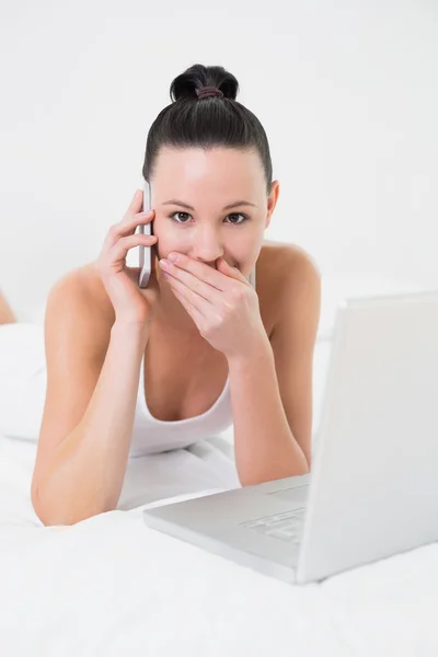 Frau mit Handy und Laptop im Bett schockiert — Stockfoto