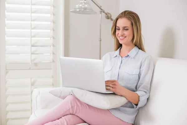 Smiling casual blonde sitting on couch using laptop — Stock Photo, Image