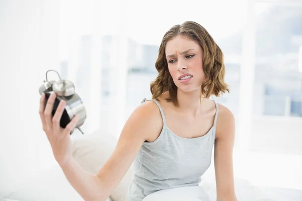 Schöne genervte Frau hält einen Wecker und sieht ihn sitzend an — Stockfoto