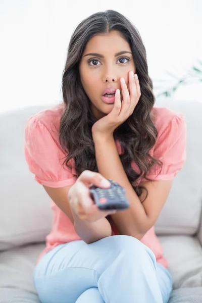 Shocked cute brunette sitting on couch holding remote — Stock Photo, Image