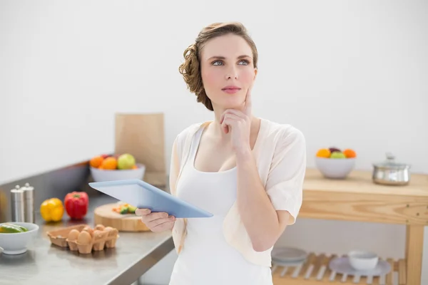 Thoughtful beautiful woman holding her tablet while standing in her kitchen — Stock Photo, Image