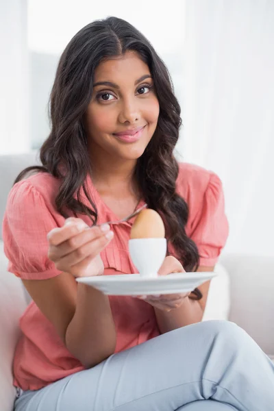 Zawartość ładny brunetka siedzi na kanapie trzymając twardych gotowane jajko — Zdjęcie stockowe