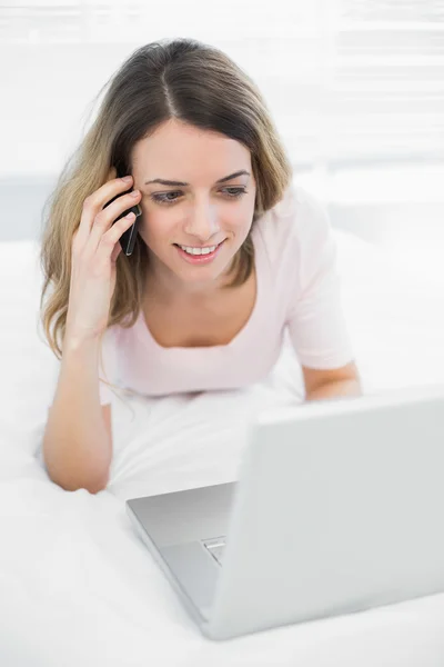 Vrolijke brunette vrouw bellen met smartphone liggend op haar bed — Stockfoto