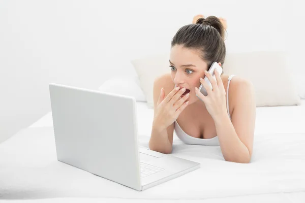 Mujer conmocionada usando el teléfono celular y el portátil en la cama — Foto de Stock