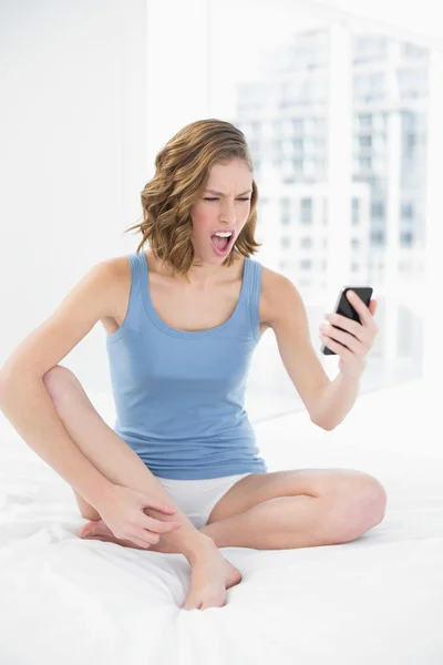 Rozzlobený krásná žena, sedící na posteli, při pohledu na její smartphone — Stock fotografie
