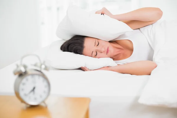 Mujer cubriendo orejas con almohada y despertador en mesa auxiliar —  Fotos de Stock