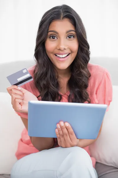 Happy cute brunette sitting on couch holding credit card and tablet — Stock Photo, Image