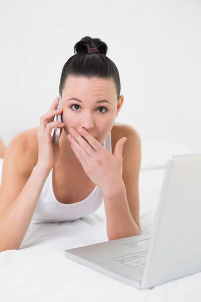 Frau mit Handy und Laptop im Bett schockiert — Stockfoto