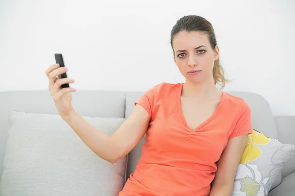 Mujer linda seria sosteniendo su teléfono inteligente sentado en el sofá — Foto de Stock