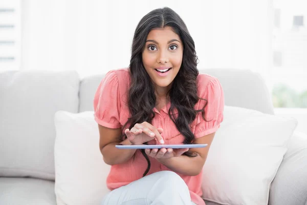 Upphetsad söt brunett sitter på soffan håller tablet — Stockfoto