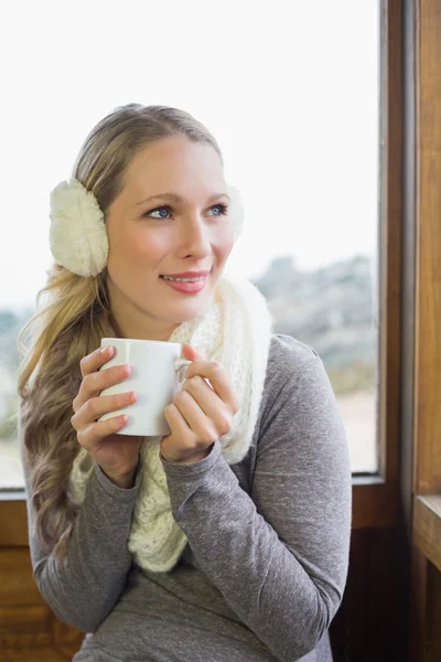 Mujer sonriente usando orejera con taza contra ventana — Foto de Stock