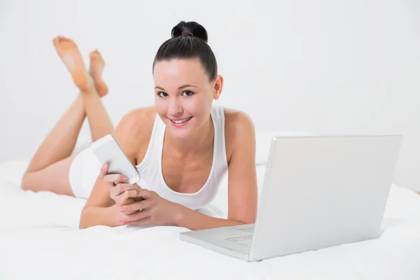 Mujer casual sonriente usando teléfono celular y portátil en la cama —  Fotos de Stock