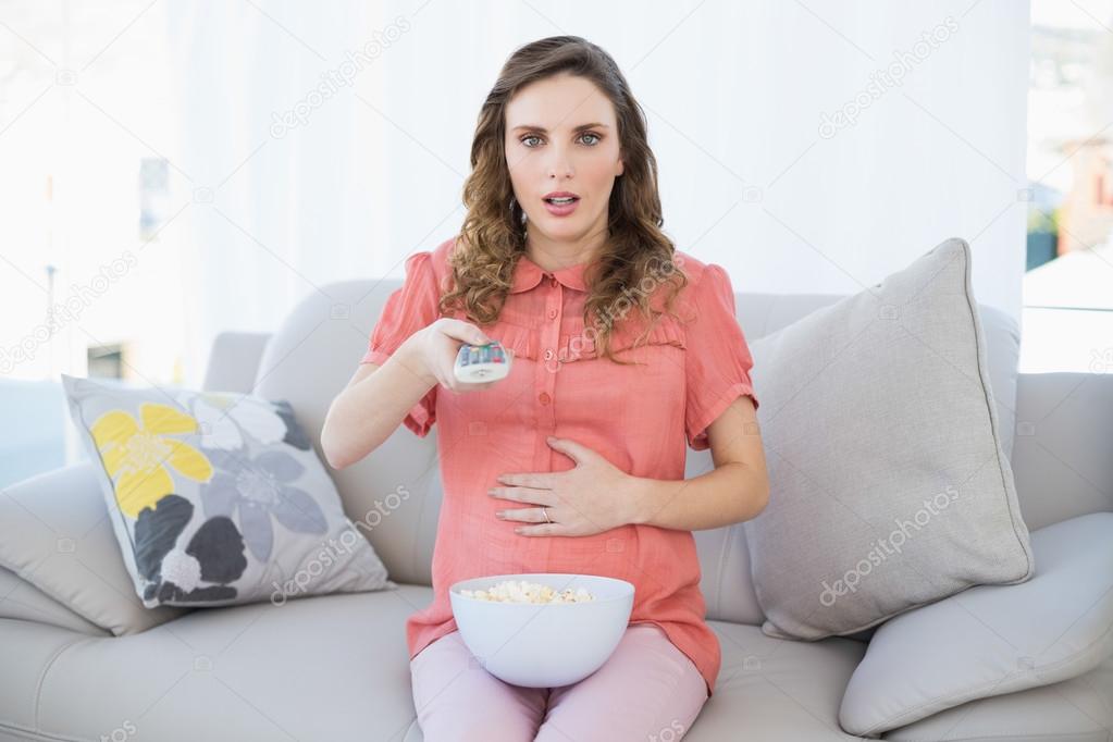 Astonished pregnant woman sitting in living room watching television at home