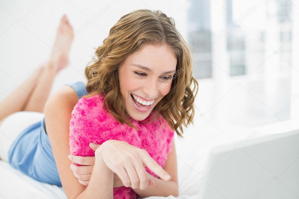 Lovely laughing woman cuddling with a heart pillow pointing at her notebook