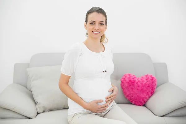 可爱的孕妇自豪地微笑着坐在沙发上的照相机 — 图库照片