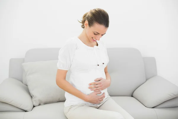 Stolze Schwangere sitzt auf Couch und berührt ihren Bauch — Stockfoto