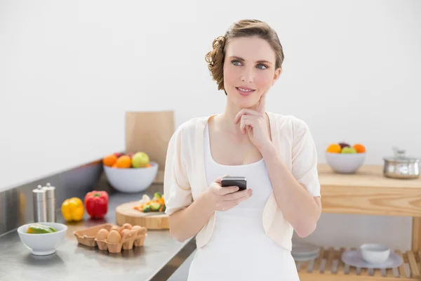 Thoughtful pretty woman holding her smartphone standing in her kitchen — Stock Photo, Image