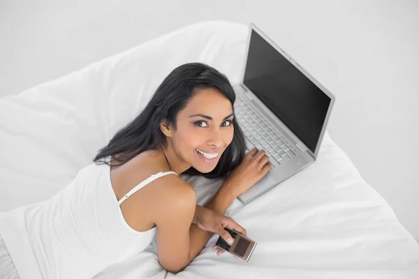 Felice donna sorridente sdraiata sul suo letto tenendo il suo telefono — Foto Stock