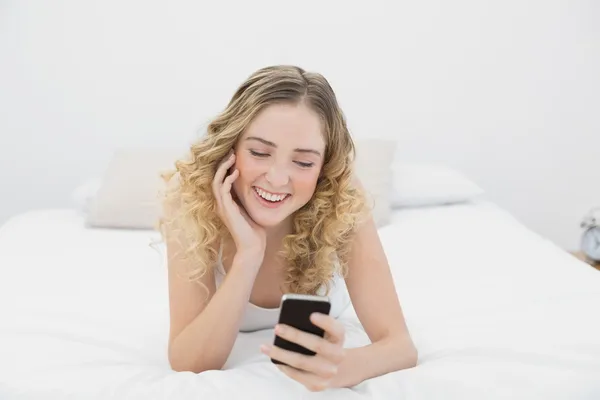 Dość uśmiechający się blondynka, leżąc na łóżku za pomocą smartphone — Zdjęcie stockowe