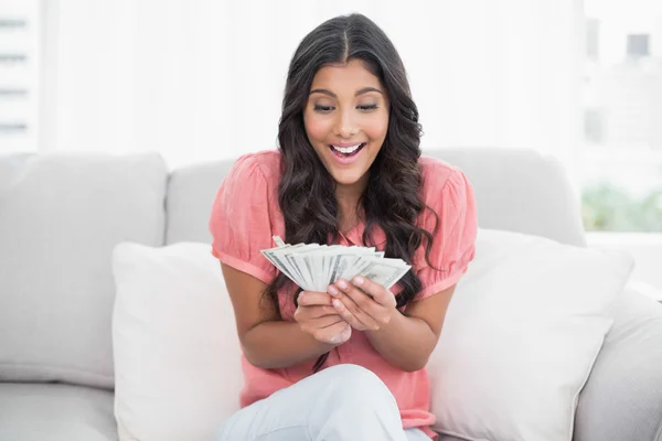 Excited cute brunette sitting on couch holding money — Stock Photo, Image