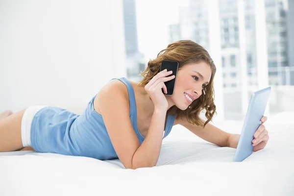 Sweet woman lying smiling on her bed holding her tablet while phoning — Stock Photo, Image