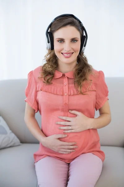 Ładny ciąży kobieta siedzi na kanapie, jednocześnie relaksując się i słuchania muzyki — Zdjęcie stockowe